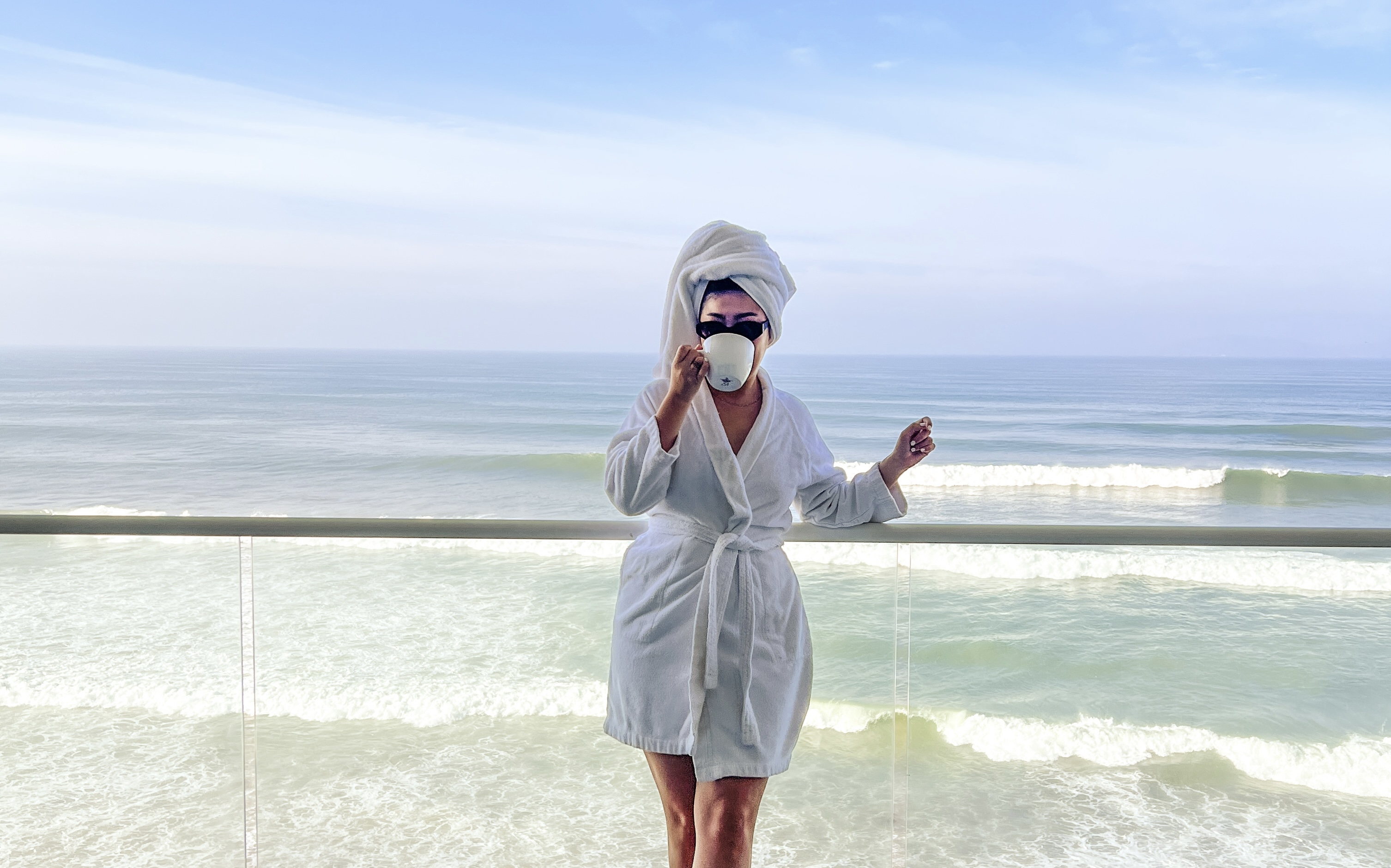 Yuna in a hair towel, bathrobe, and sunglasses sipping a cup of coffee while standing on a balcony with the ocean in the background
